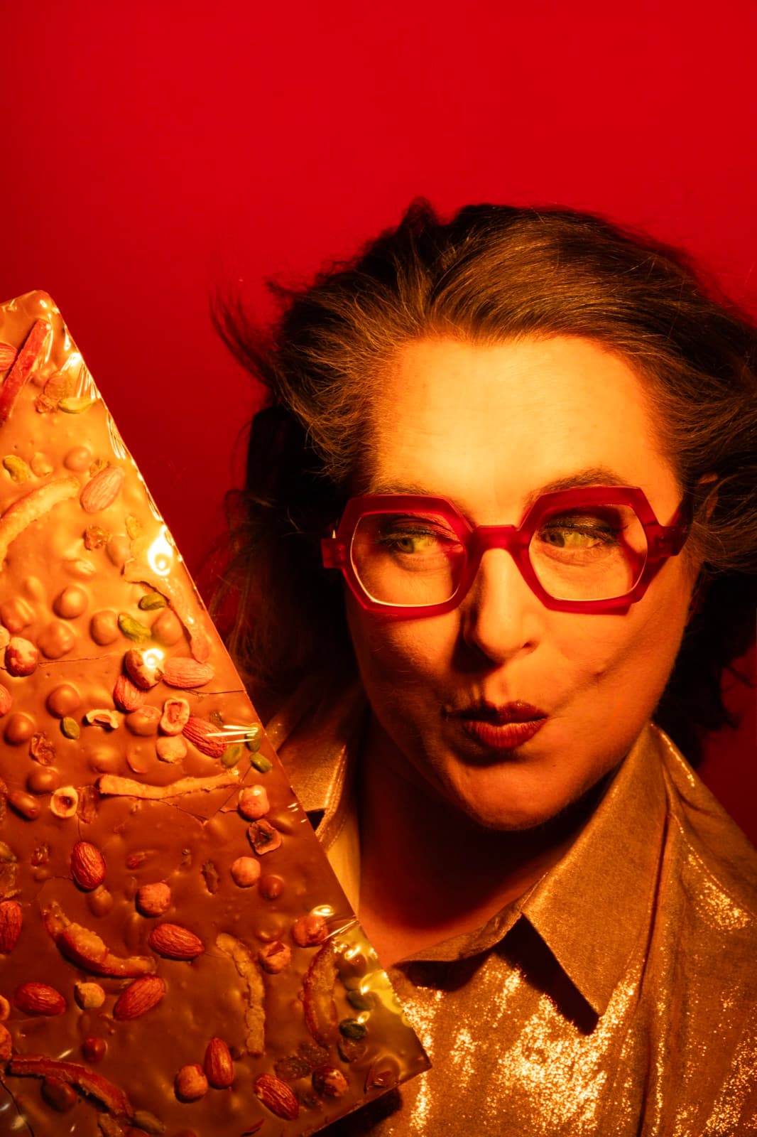 Mélanie Coudert, lunettes signature et veste dorée, présentant une tablette de chocolat d'exception sur fond rouge — Volcanique & Pimenté